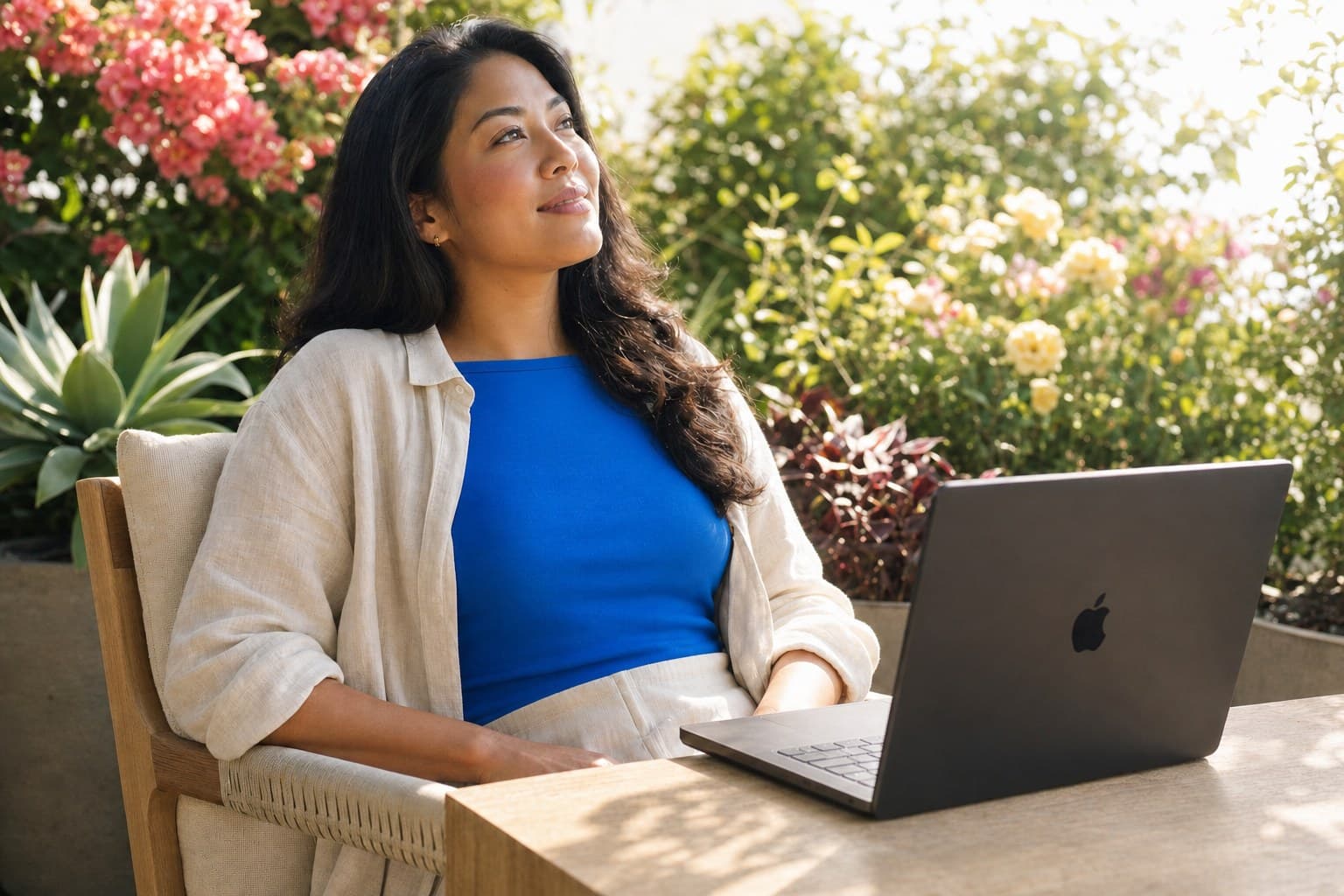 A woman in her early thirties sitting back easily in a chair on a flowering garden terrace, wearing bright blue under an open cream overshirt, her face lifted and turned aside with a small content smile, her hands away from the keyboard while a Space Black MacBook Pro sits open beside her